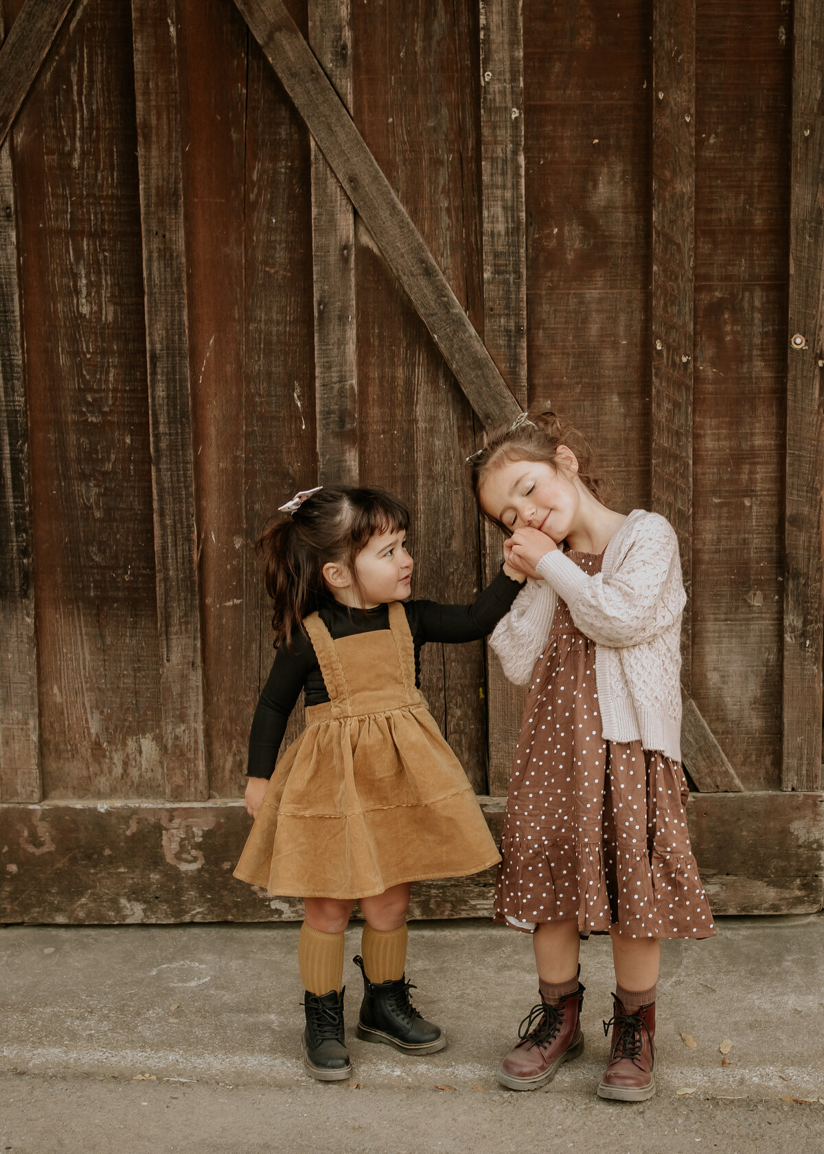 Sister portrait in front of barn. Older sister holds younger sister's hand to cheek.