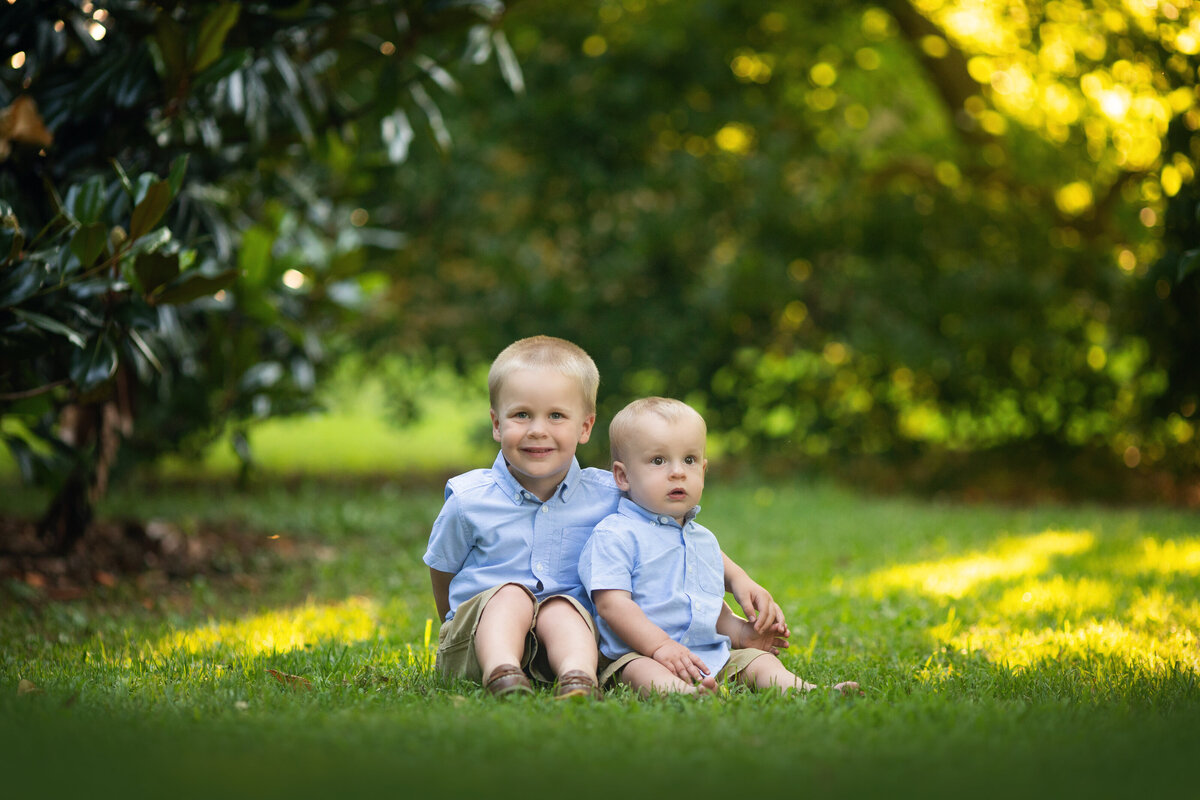 A toddler boy in a blue shirt sits in a park with in younger brother in a matching shirt