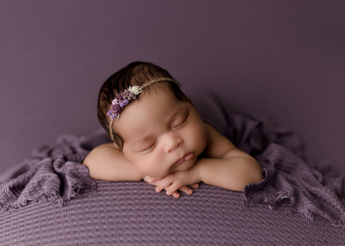 Newborn photoshoot in Boca Raton studio with top photographer. Baby is resting on a purple textured stretch fabric, laying on her belly. Baby's hands are folded under her chin and is resting her head on her left arm. Baby is wearing a delicate purple floral headband and a matching textured purple blanket is draped over her back.