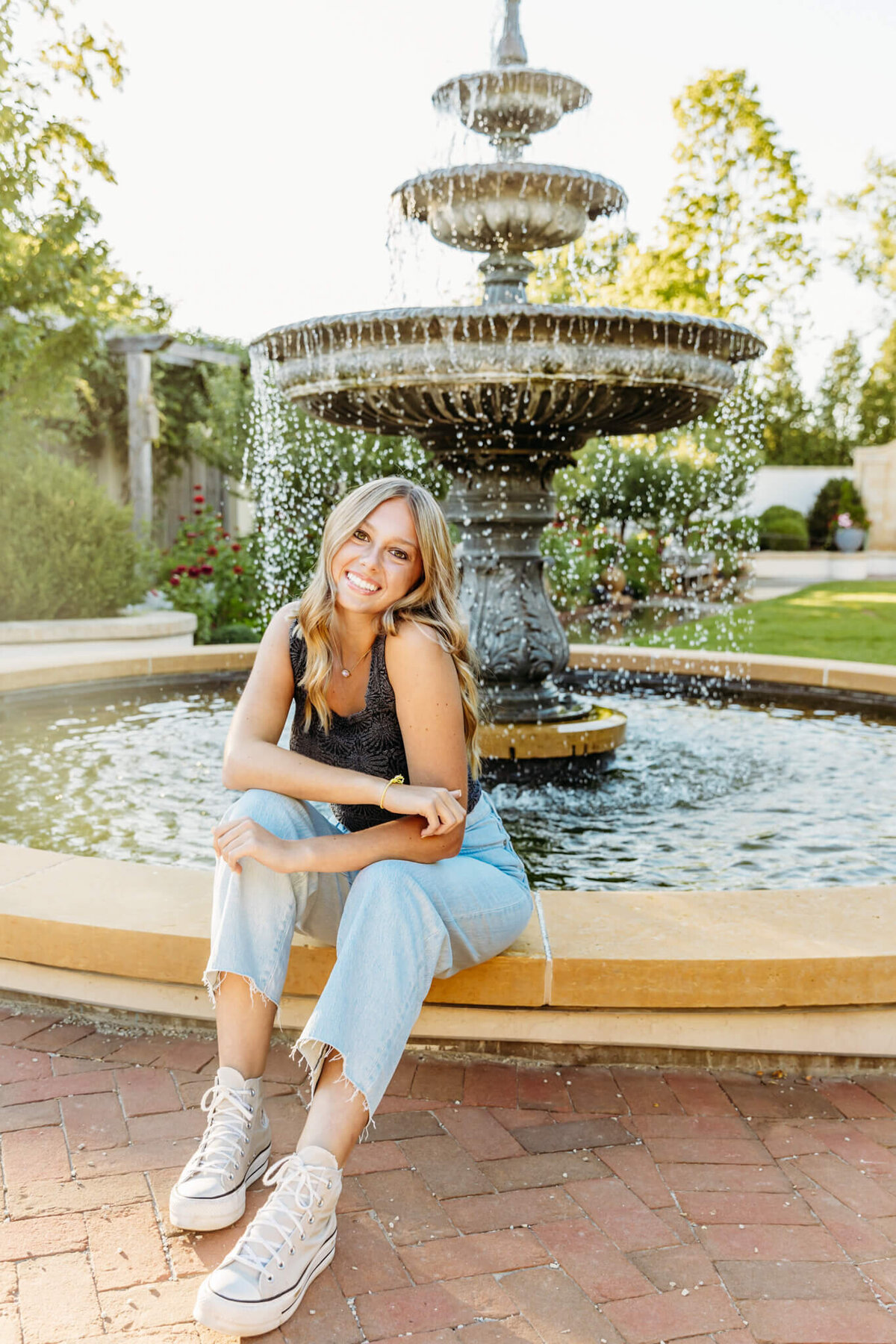 high school girl in grey top and jeans sitting  by a fountain and leaning forward while resting her elbows on her knees captured by Ashley Kalbus Photography