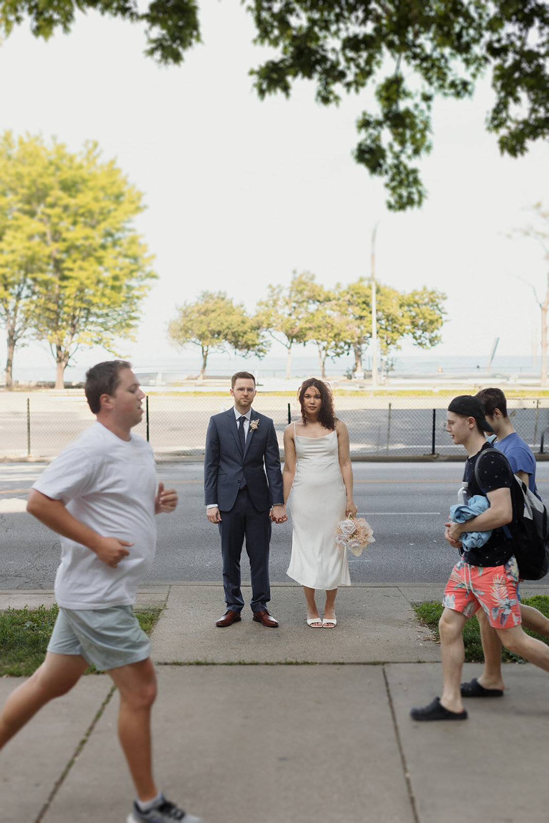 Just Married photo session couple stands along Lake Shore Drive and holds hands with runners passing by in either direction.