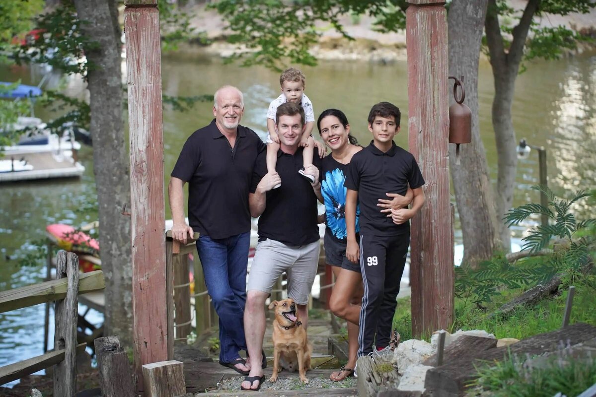 A family of five smiling with a dog in front of a lake.