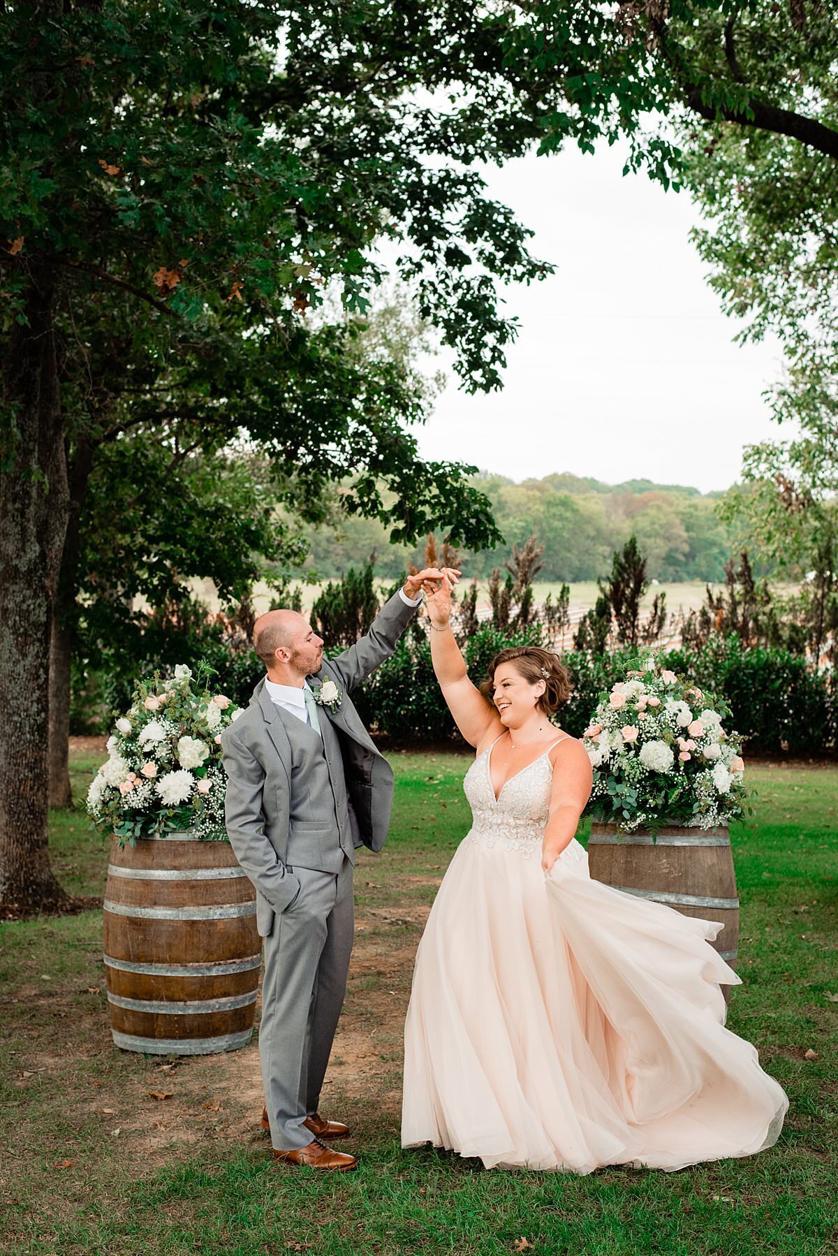 Bride wearing a tulle skirt ballgown in a soft blush twirling with her husband in front of wine barrels with large flower arrangements on top of them