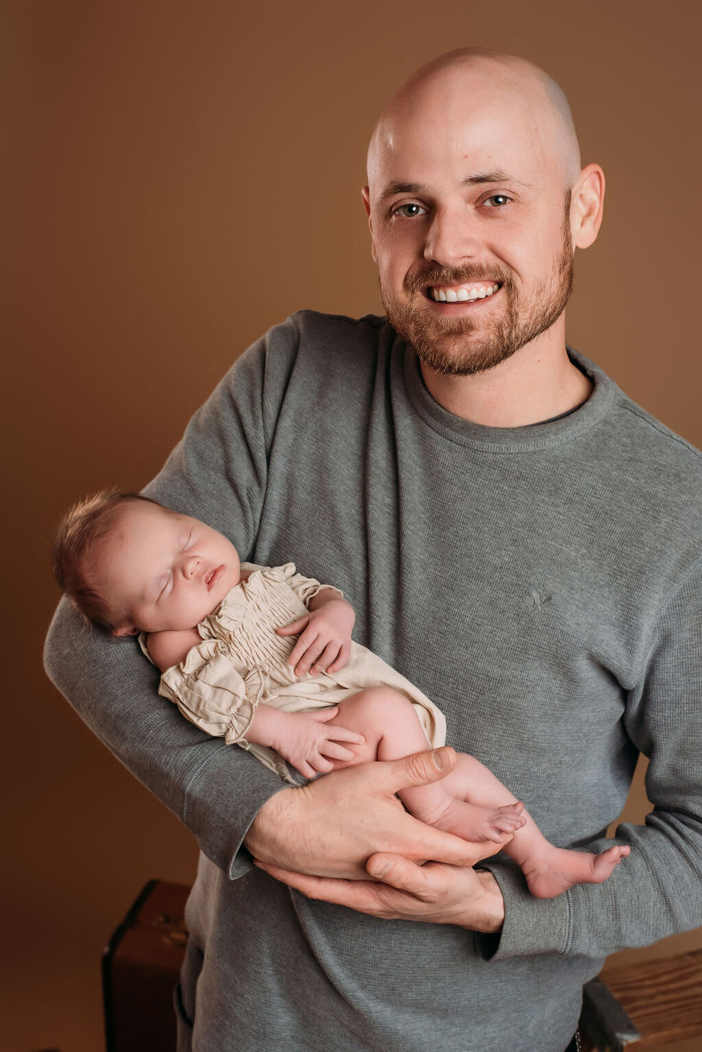 A man in gray sits in front of a neutral-toned backdrop holding a newborn baby in a beige outfit, captured by a photographer in Bemidji, Minnesota.