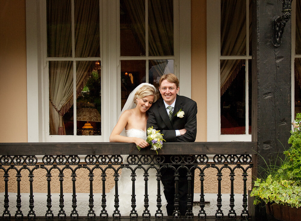 Bride with blonde hair, wearing a princess style wedding dress, holding a white flower bouquet, relaxing with her groom on the veranda of the Muckross Park Hotel