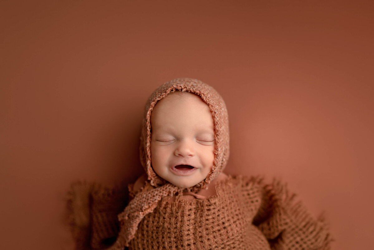 Newborn Baby girl swaddled in rust blanket and bonnet, laying on a rust colored blanket smiling at the camera.