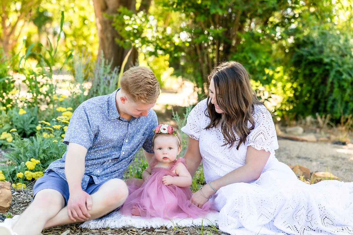 Family of 3 posed in a gardern.  Dad is wearing a blue shirt and shorts, mom is wearing a white eyelet dress and both are looking at the toddler.  Toddler girl is wearing a mauve lace dress with a flower crown and looking at the camera