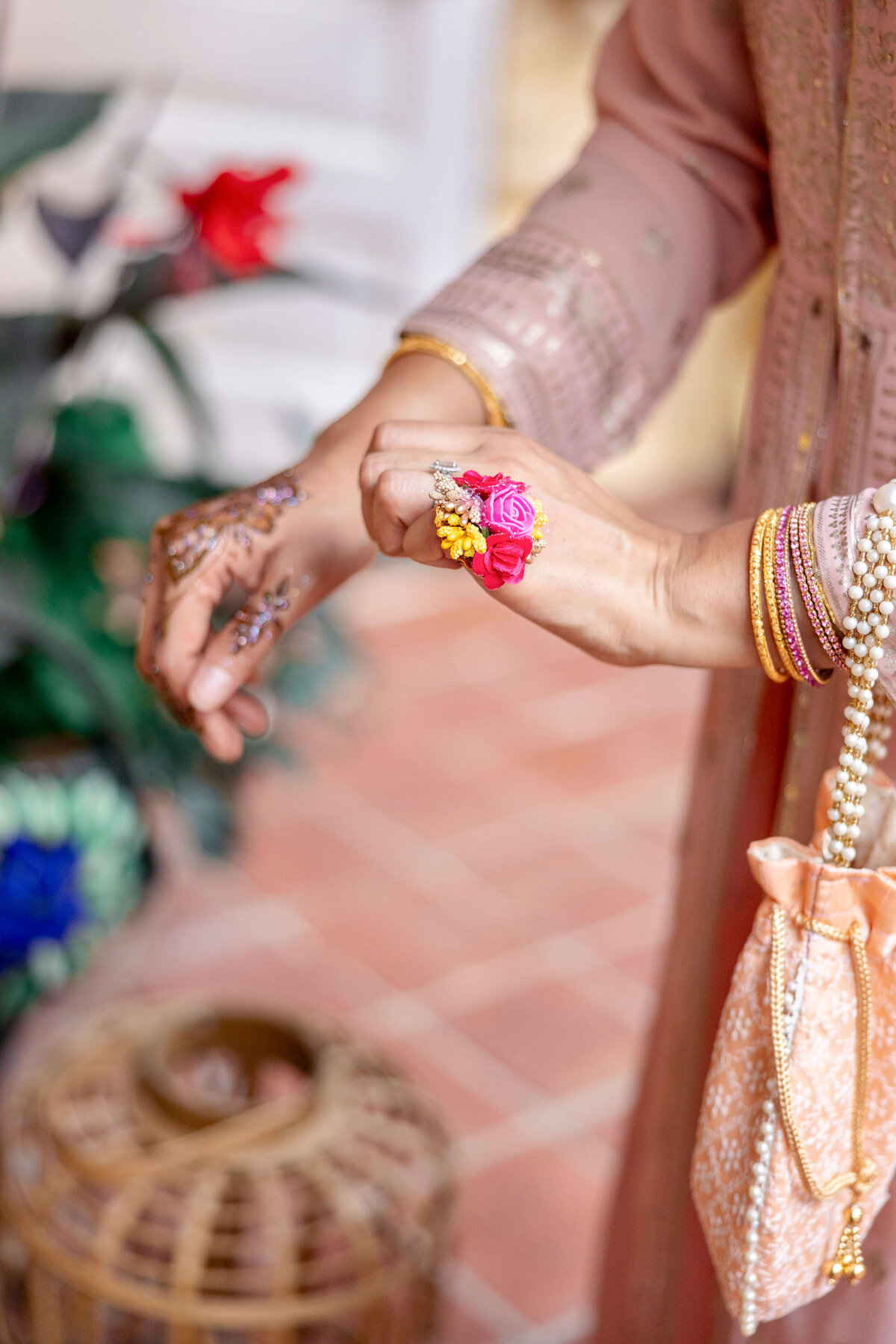 A person wearing a traditional pink outfit decorates their hand with intricate henna designs. They have colorful floral jewelry and hold a peach embroidered clutch. In the background, there are plants and a woven basket.