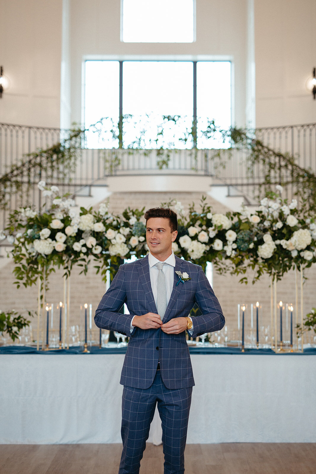 Groom in a blue suit buttons his jacket in front of a long rectangular table with lit candles and white florals.