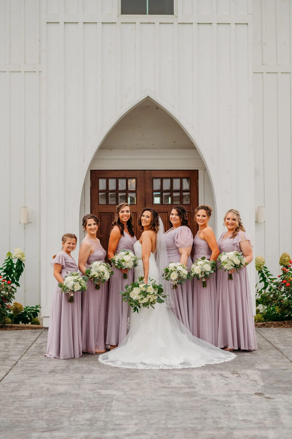 Photo of a bride and her bridesmaids, and lavender, in front of a white building and wooden doors