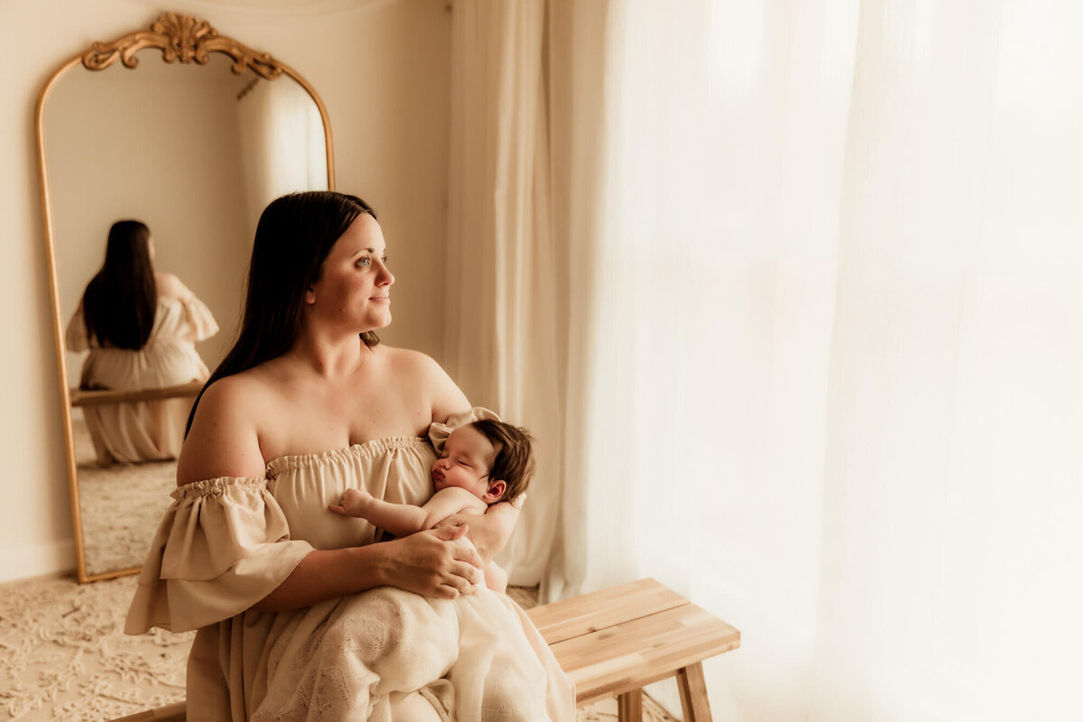 Mother looks the window while holding her sleeping baby girl.