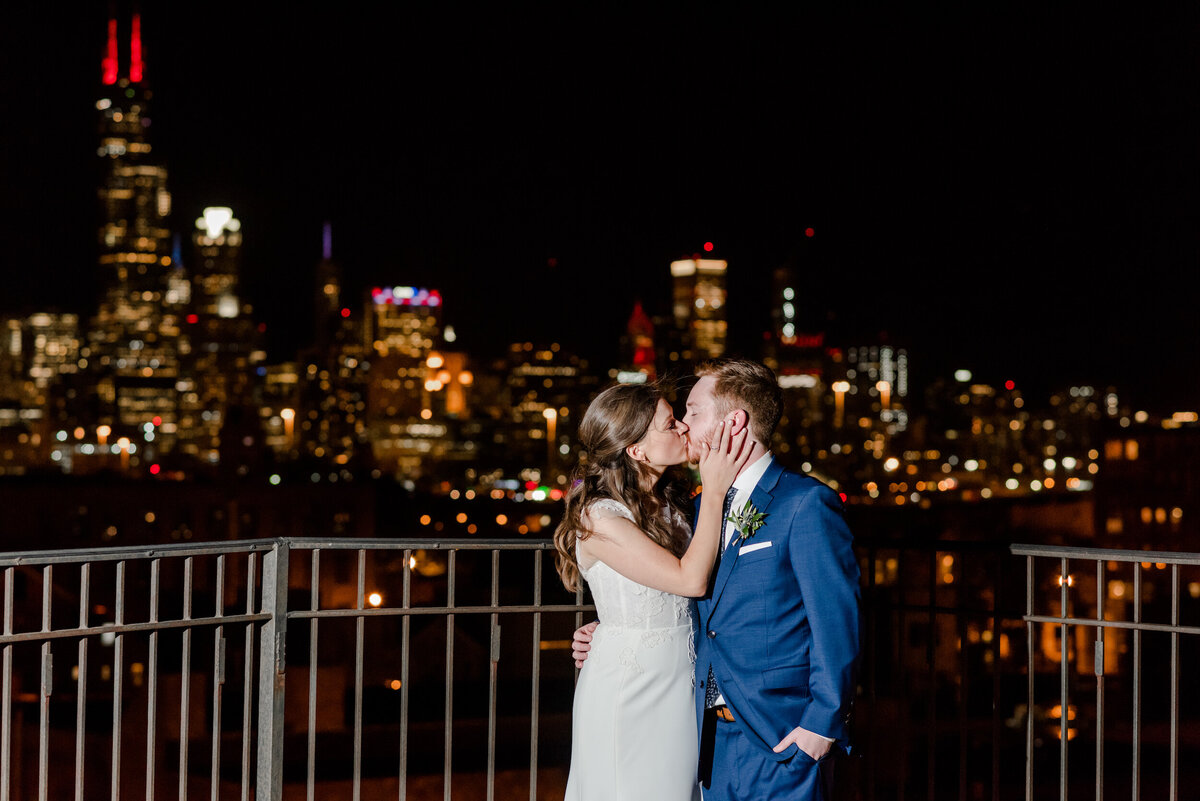 Bride and groom kiss in front of the Chicago skyline at night on the rooftop at Lacuna Lofts