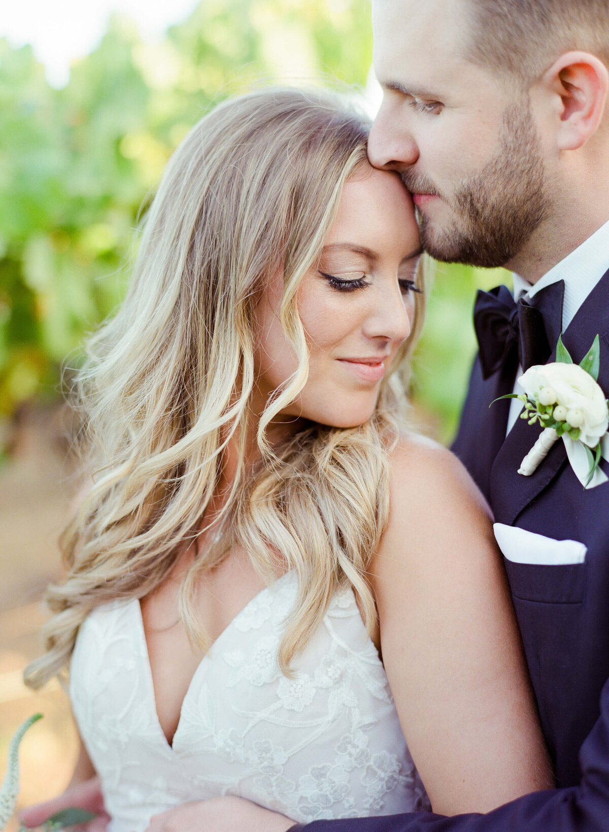 Beautiful bride in Deep V-cut sleeveless wedding gown gets kissed on the forehead by her beloved husband. Vineyard wedding photography in Wine Country by Robin Jolin.
