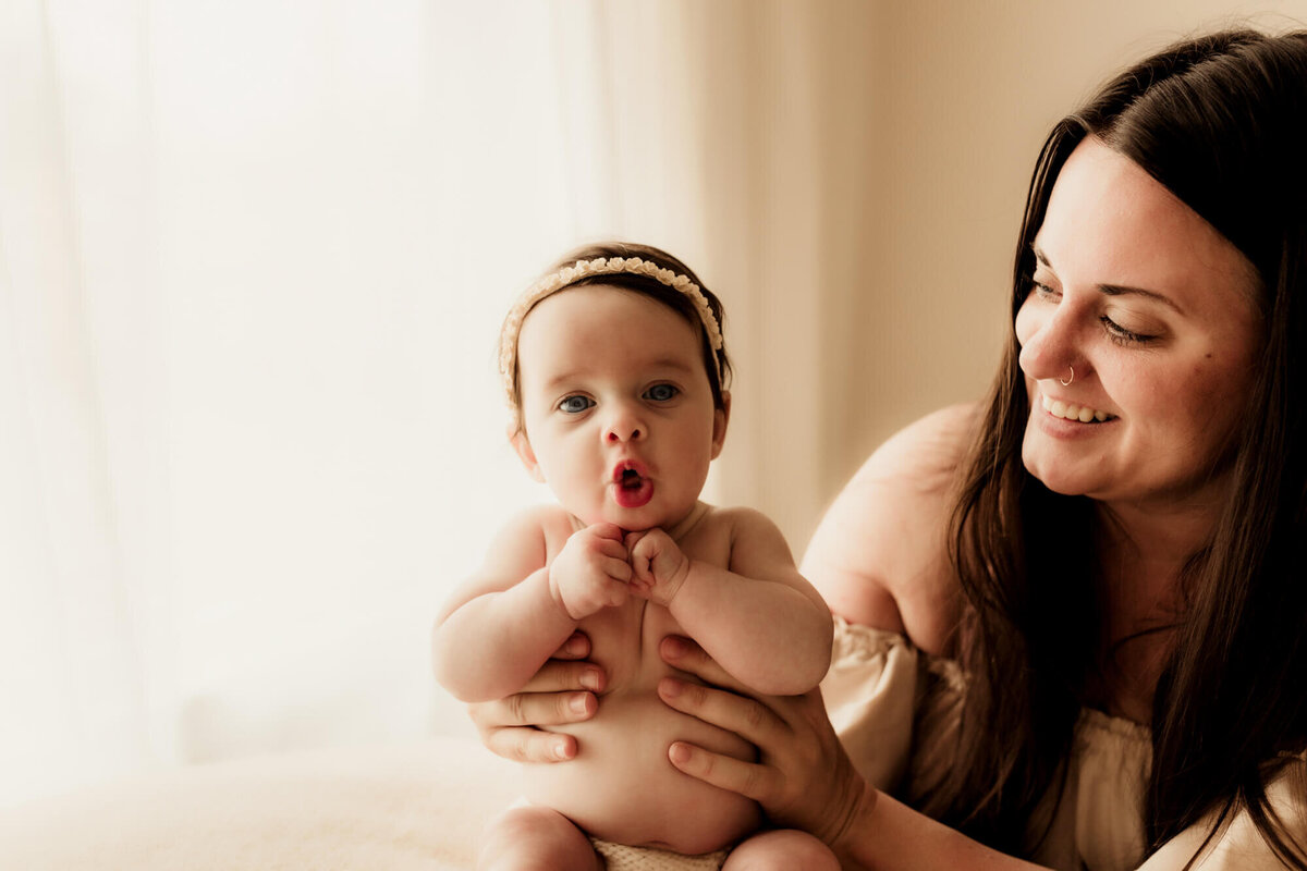 Baby girl makes a cute expression as her mother smiles at her near a window.