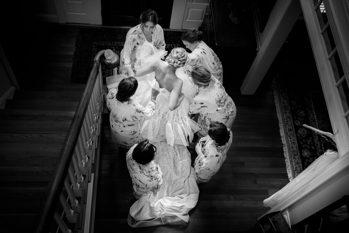 Overhead black and white photo of a bride surrounded by bridesmaids in robes, helping her get ready for the wedding.