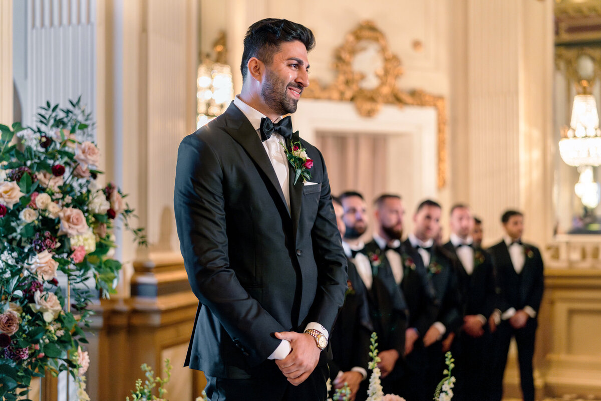 A groom in a black tuxedo stands smiling at the altar of an elegant venue adorned with floral arrangements. Behind him, groomsmen in matching suits stand in alignment, and ornate chandeliers illuminate the scene.