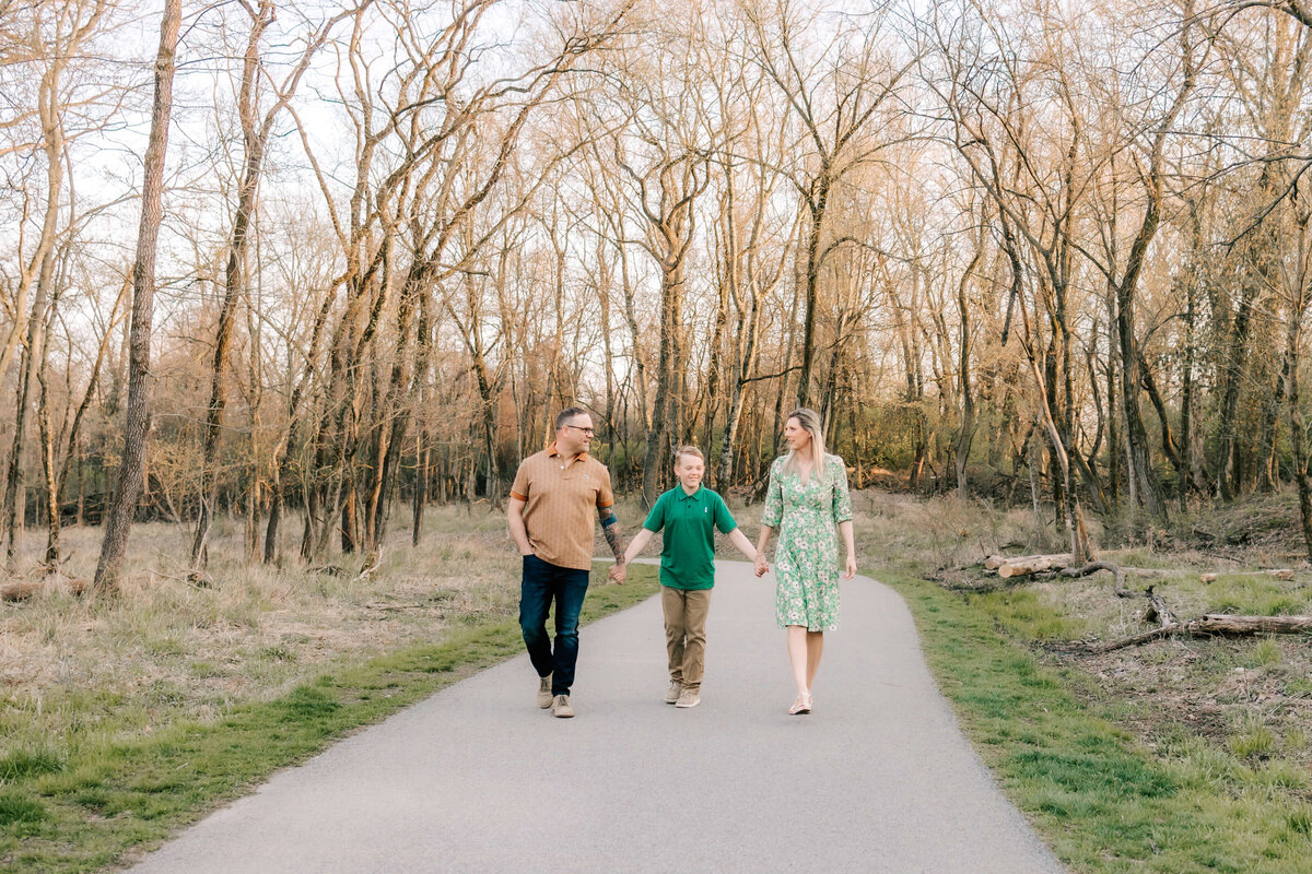 family of 3 walking holding hands and looking at each other smiling on a paved pathway surrounded by trees