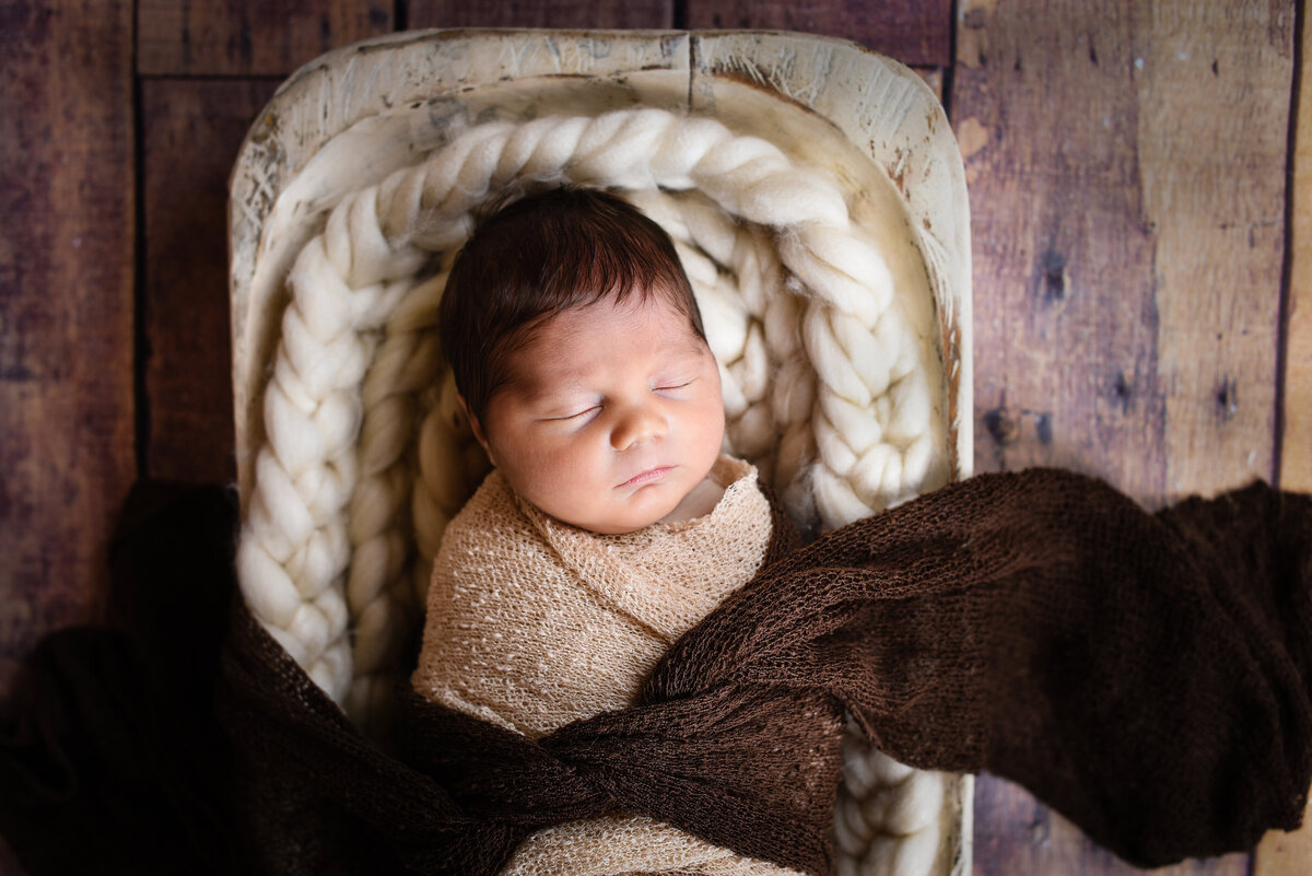 Beautiful Mississippi newborn photography: Boy wrapped in ivory with head of dark hair sleeps in dough bowl on wood floor