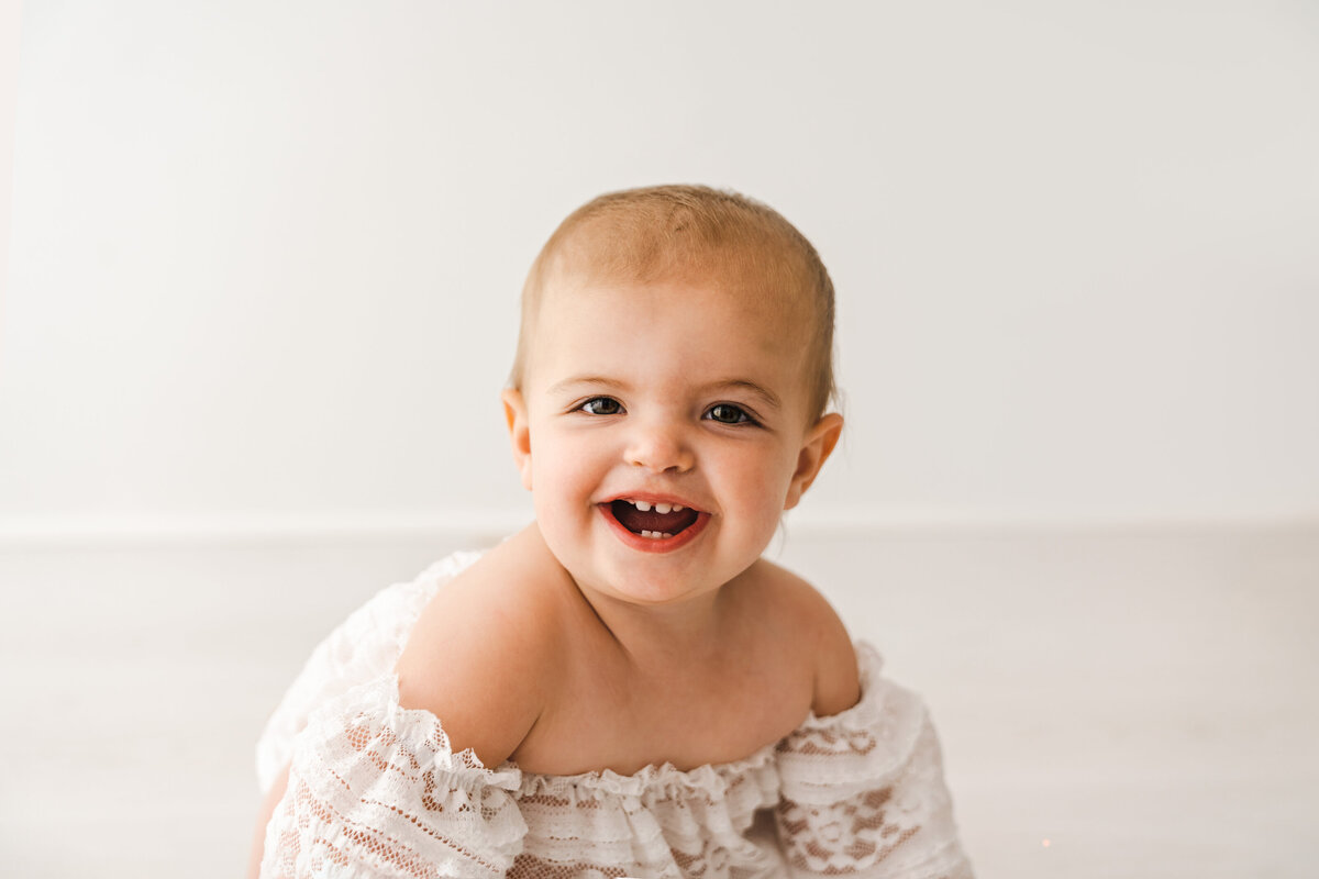 Joyful baby with a bright smile showing tiny teeth, dressed in a lacy outfit, against a clean, white background. Taken by Fig and Olive Photography, Minneapolis Baby Photographer.