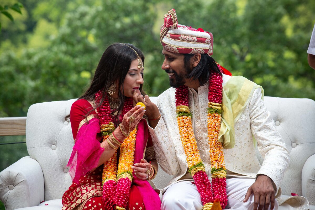 A bride in a red saree and a groom in a white and yellow outfit sitting together and smiling, amidst a green park setting, during a traditional Iowa wedding ceremony.