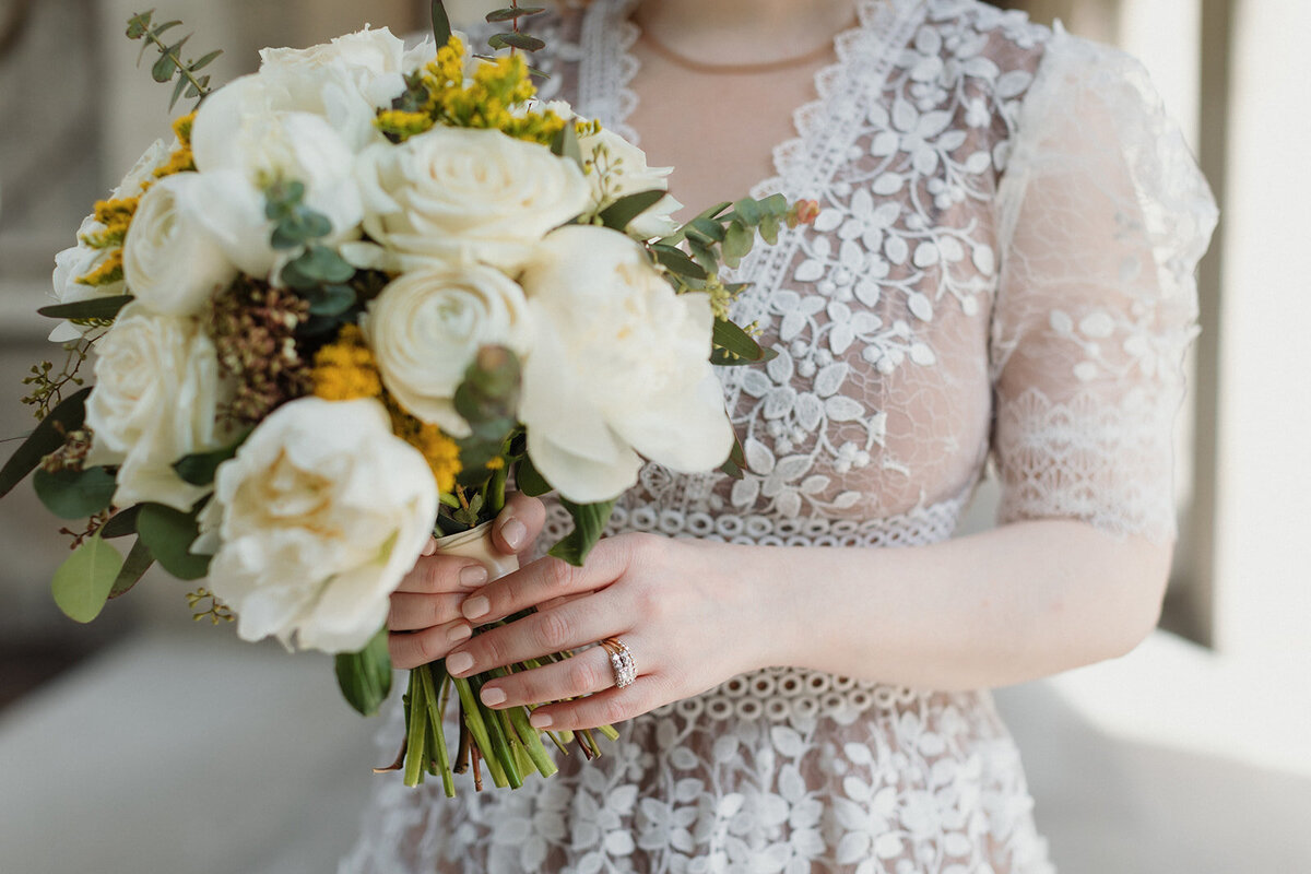 Very close up photo of bride carrying a yellow and white bouquet, focus is on her ring. Her dress is beautifully detailed with lace.