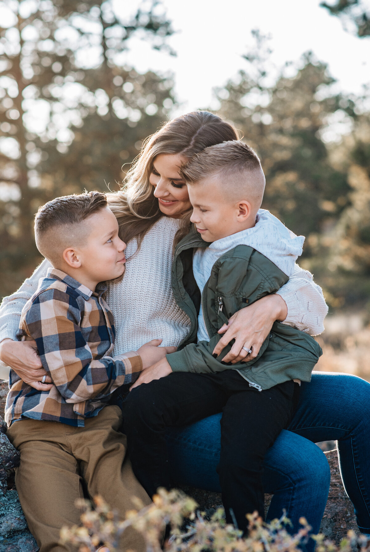 denver family photographers photographs mother sitting with her two young children who are sitting on her lap and smiling at her as the sun sets in the distance of their fall family pictures