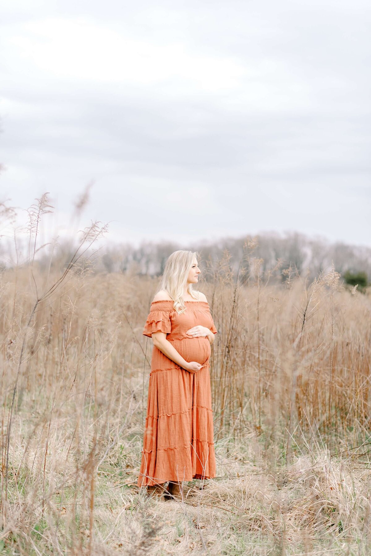 A beautiful pregnant woman poses in a golden field during the wintertime  wearing a flowy orange dress.  Captured by Charlotte Maternity Photographer, Melissa Mayrie Photography