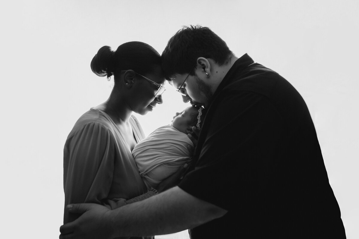 Newborn family portrait of dad kissing mom's forehead and mom kissing baby's nose at Marietta, GA newborn photography studio with mom, dad and baby girl wearing blush pink, cream and black. Black and white portrait.