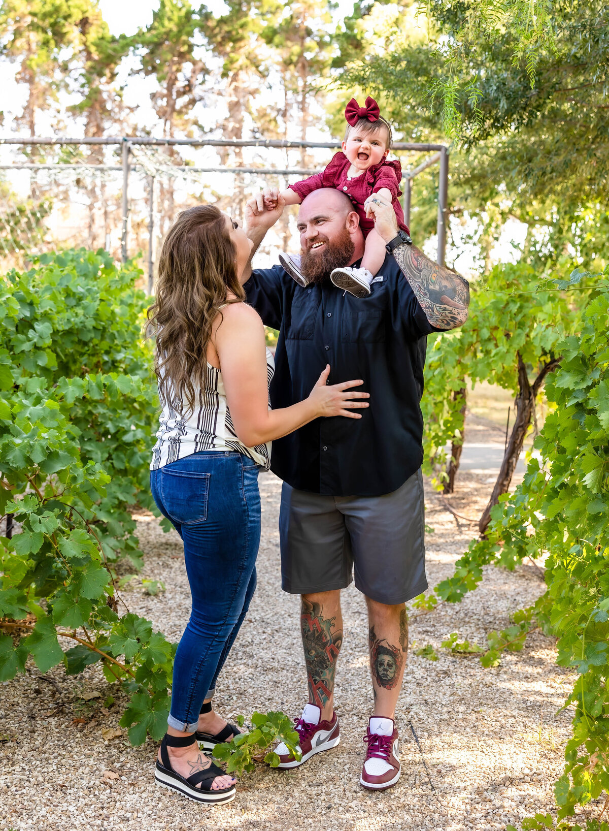 Family is posed in a vineyard with mom and dad looking at each other and baby is looking at the camera.  Dad is wearing a black shirt and shorts and mom is wearing a striped sleeveless shirt with jeans and sandals.  Baby is in a maroon romper with a matching hairbow