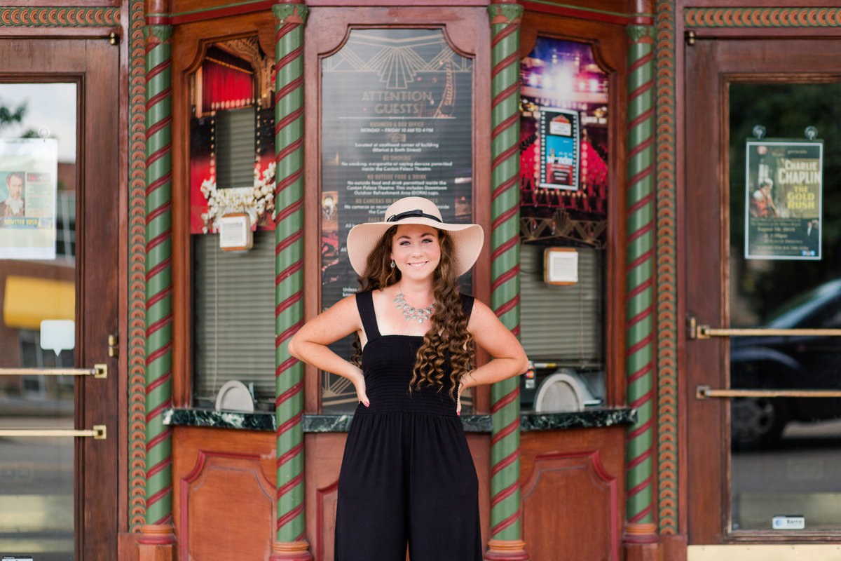 high school senior girl wearing black jumper with high heel sandels, also wearing a tan hat standing with hands on hips in front of Canton palace theater in Canton Ohio photographed by Jamie Lynette Photography Canton Senior Photographer