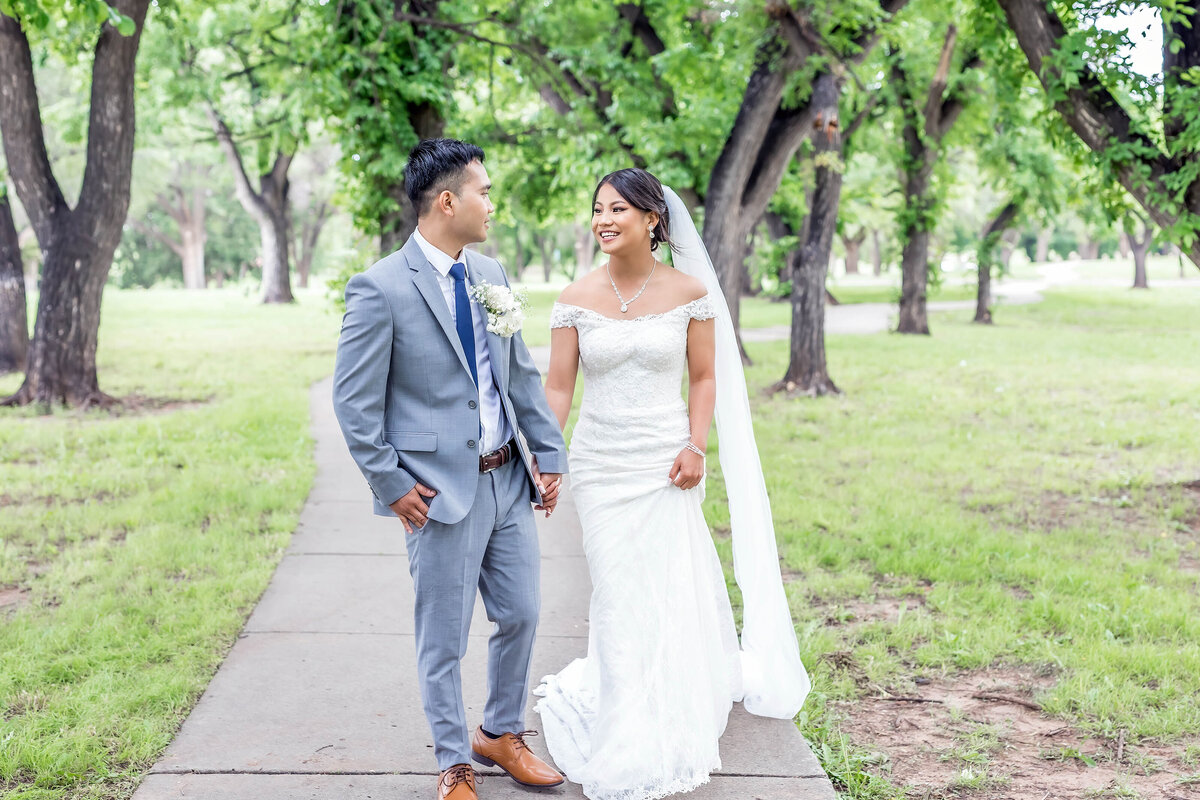 Burma couple walking and looking at each other.  Groom is wearing a gray suit and bride is wearing a white wedding dress with a long veil.