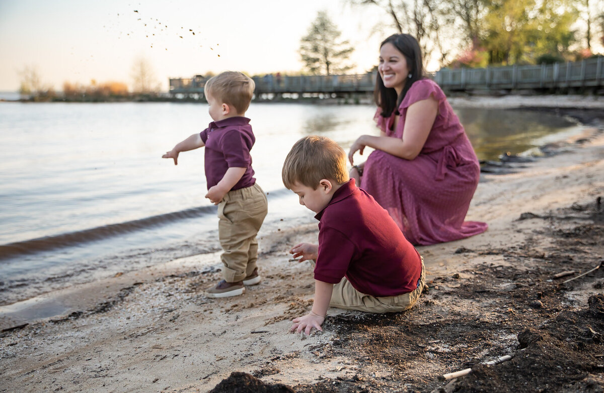 A child throws sand while his Moother smiles at him. Their brother plays with the sand.  This is a candid photo during their lifestyle family photos in Harford County, Maryland