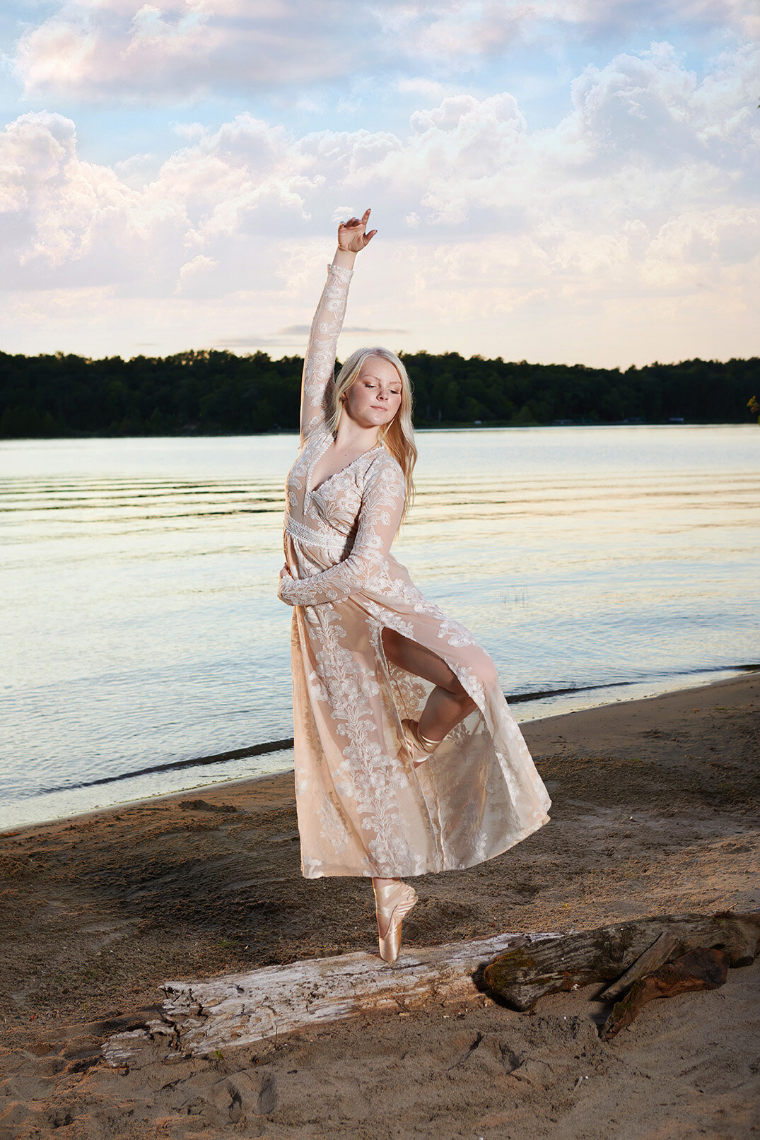 Blonde haired high school senior girl strikes a ballerina pose on dead tree laying on the beach Leech Lake wearing long pink lace dress.