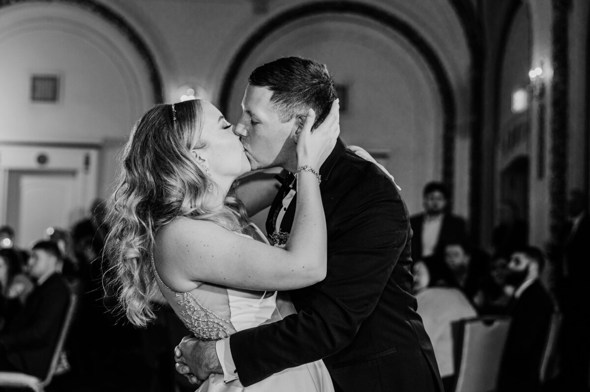 A black and white photo of a bride and groom kissing passionately during their park farm winery wedding, with guests in the background.