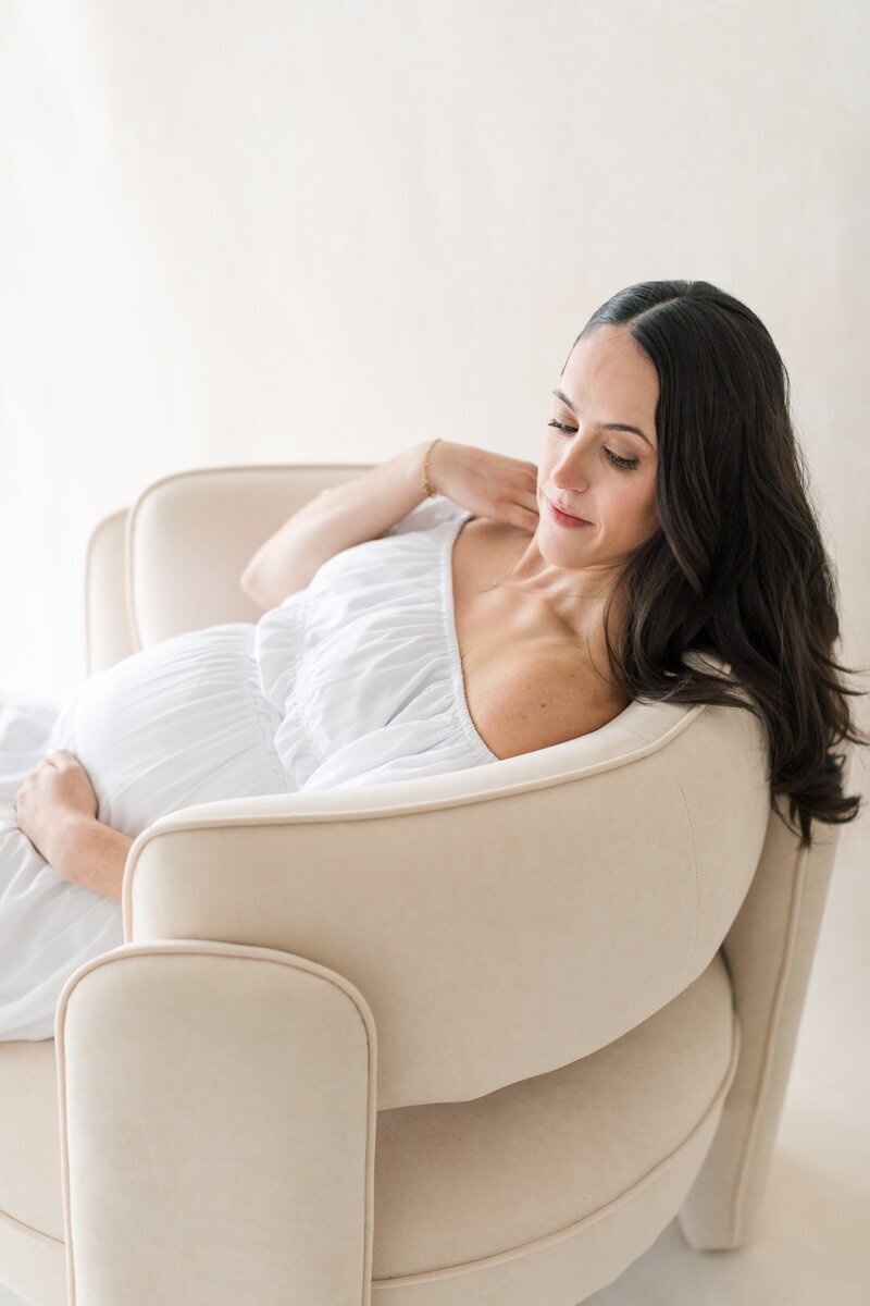 A pregnant woman wearing a white off the shoulder dress relaxes back in a chair and holds her pregnant belly as she gazes down at it during her Lawrenceville NJ maternity photography sessions