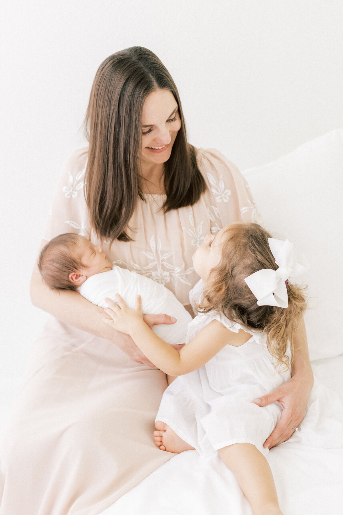 mom in pink dress cuddling her toddler girl with a big white bow while holding her newborn baby boy during her photo session