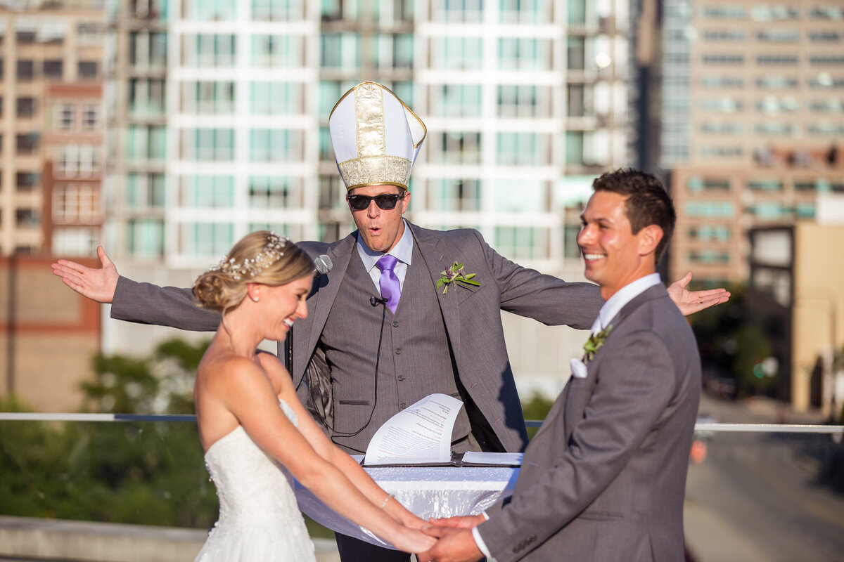 A friend wearing pope's hat and black sun glasses officiates a wedding of a couple on a Chicago rooftop.