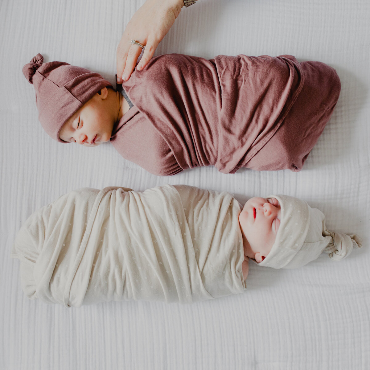 Overhead photo of baby in swaddle. Two babies actually! One twin is in a mauve swaddle and the other is in taupe. The mom's hand reaches in from the top of the photo and rests on one of the babies.