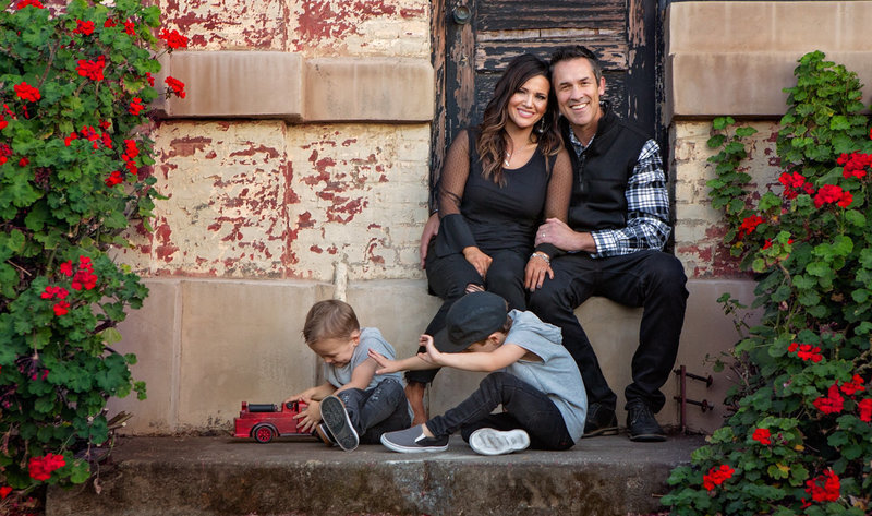 Sky 9 Studio | Professional photo of family with two boys playing with fire truck toy outside of brick building with black, grey, tones against brick wall with border of red flowers