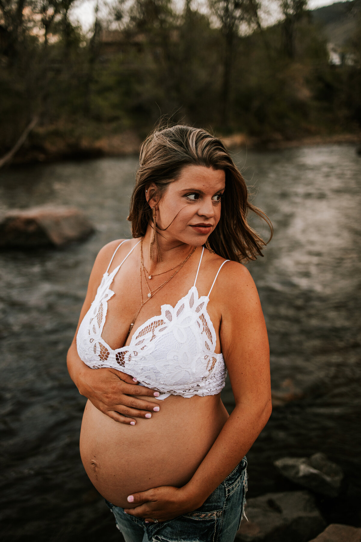 pregnant mom wears a white bralette and jeans while standing in a river in denver colorado