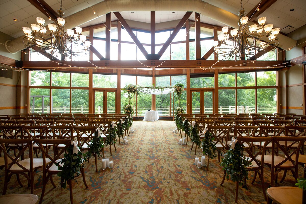 Elegant Iowa wedding venue interior, featuring rows of wooden chairs on either side facing an aisle leading to a small altar, decorated with white flowers and large windows in the background.