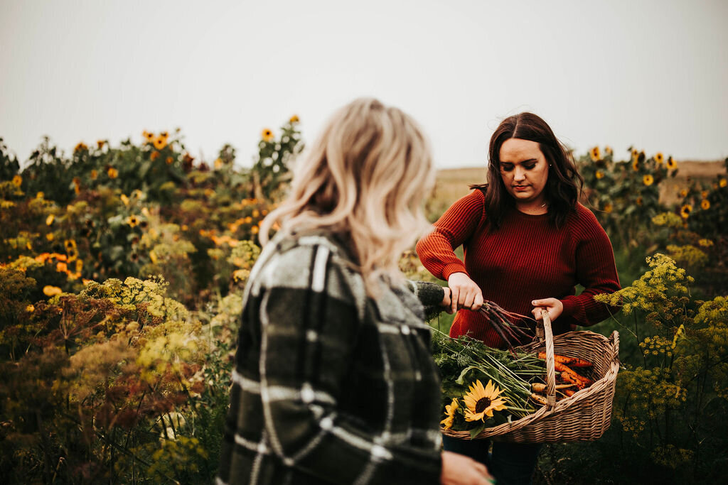 two women standing in a garden putting veggies in a basket