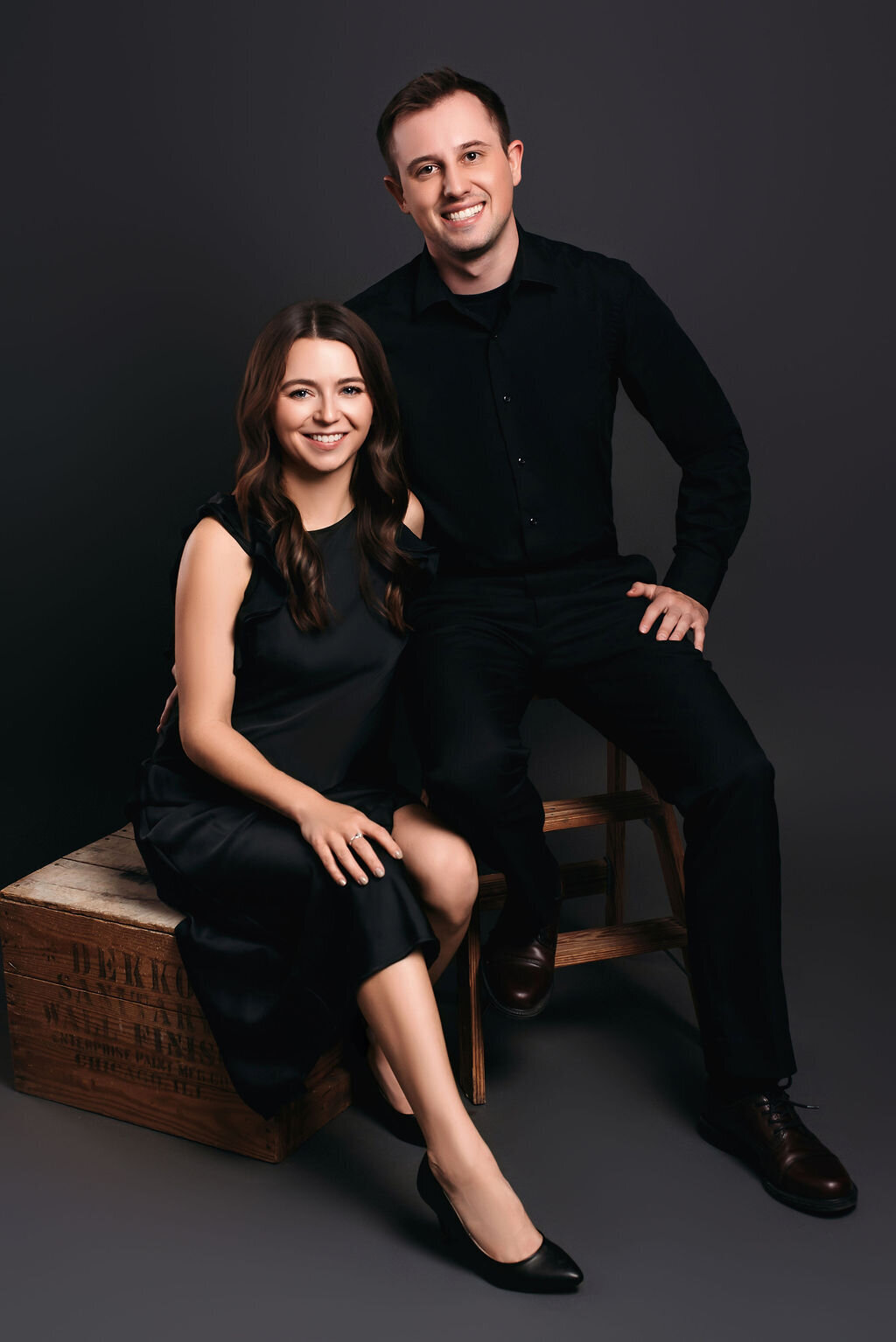 A young couple sits on rustic wooden boxes and ladders in front of a gray-toned backdrop––A woman in a black dress and heels and a man in a black dress shirt and pants, captured by a photographer in Bemidji, Minnesota.