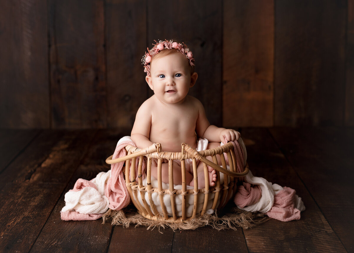 Baby girl 12-month photoshoot at West Palm Beach Lake Worth photo studio. Baby is wearing a floral headband and sitting in basket draped in pink and white linen. Baby is smiling at the camera. Basket is on a barn wood infinity backdrop.