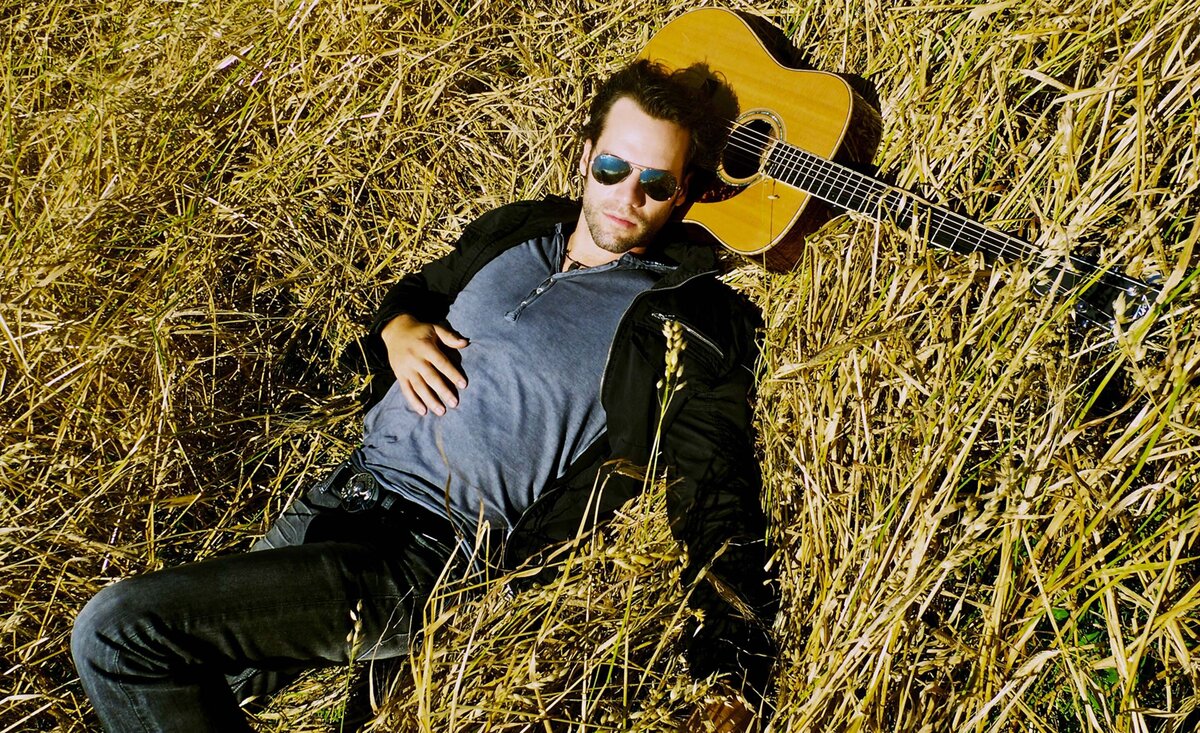 Male musician photo Chad Brownlee wearing sunglasses black coat blue shirt lying down head against guitar surrounded with long yellow grass
