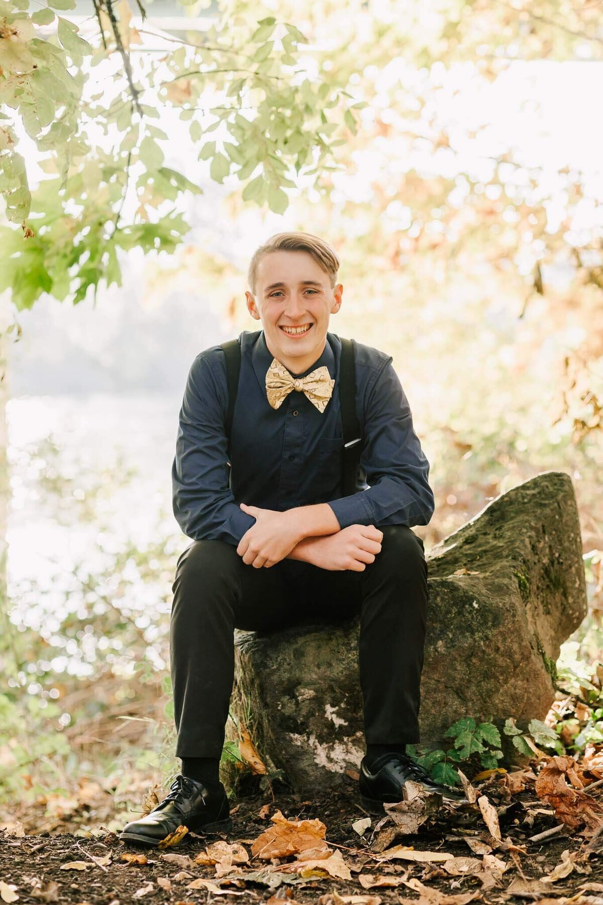 A high school senior sits on a rock at Takena Landing Park in Albany, OR. He's wearing a blue shirt and gold bowtie with black slacks and suspenders