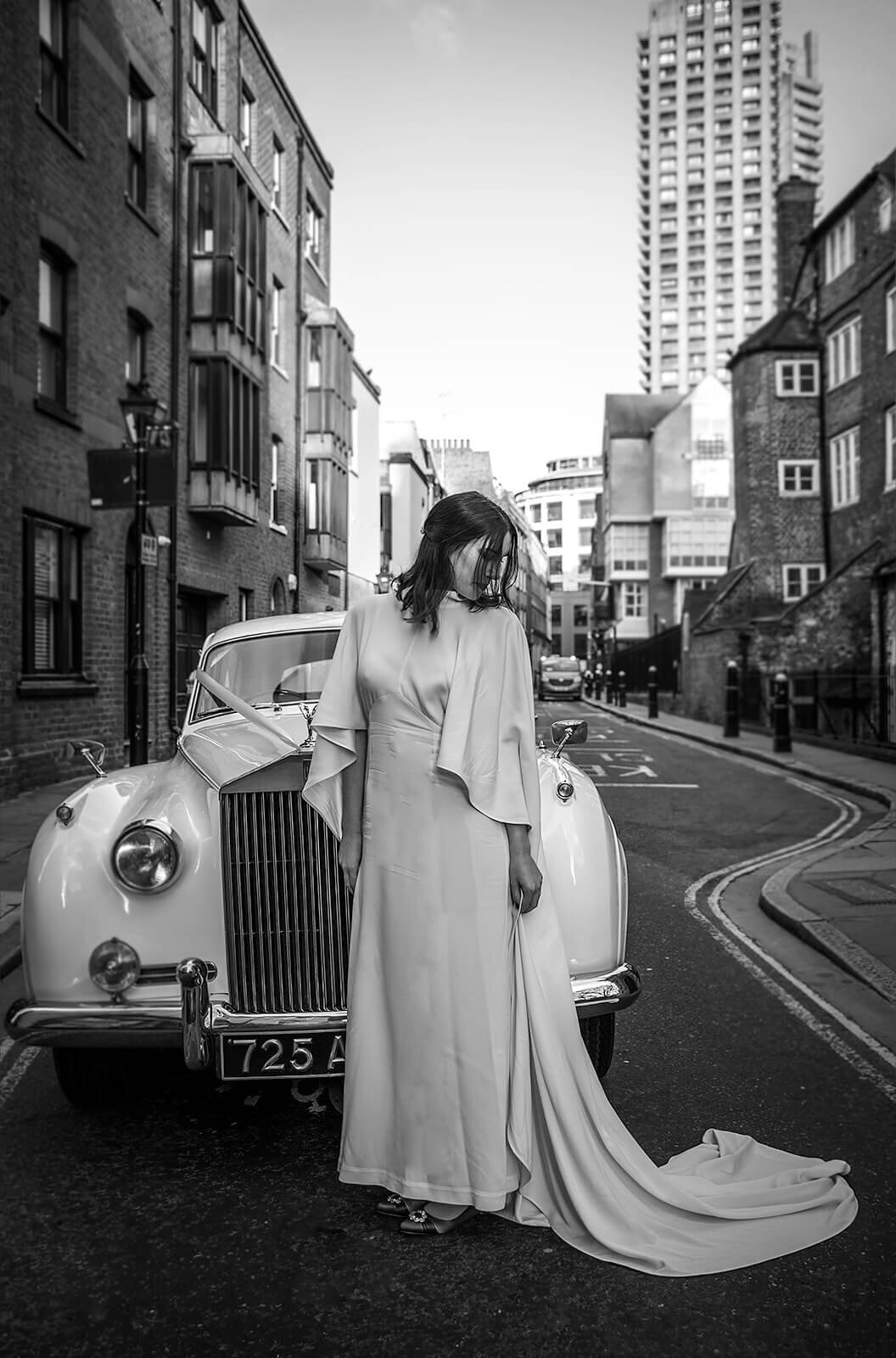 Bridal in caped dress looking down.  Standing next to rolls royce in East London street