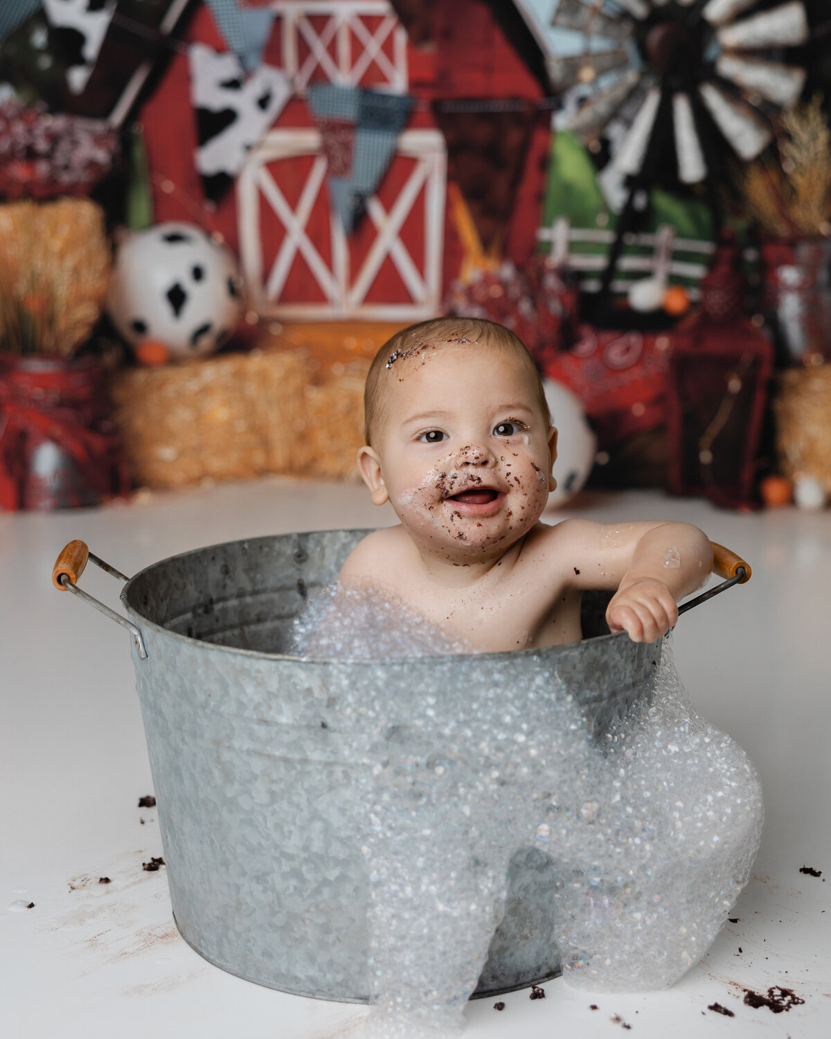 Smiling baby in a tin tub bubble bath surrounded by barnyard decor, soaking after his messy first birthday cake smash.
Photograph by Yucaipa cake smash photographer Kaitlyn Dawn Photography.