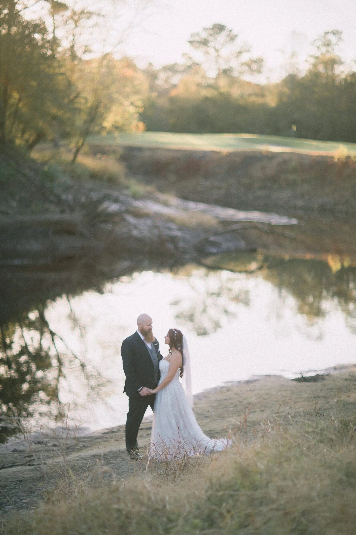 A bride and groom stand by a serene river, enveloped in a soft haze, with a picturesque landscape in the background.