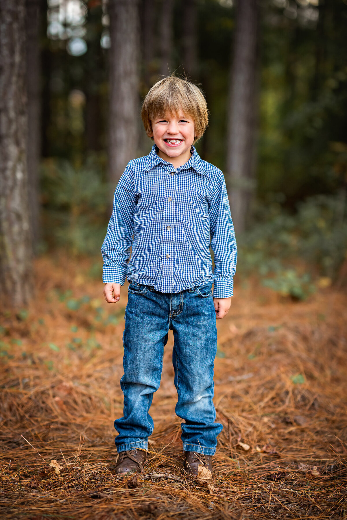 A young boy in a blue shirt is standing in the woods and smiling at the camera.